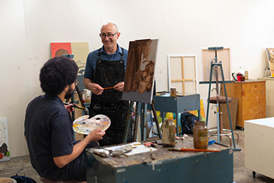 Student painting in art class with professor looking on.