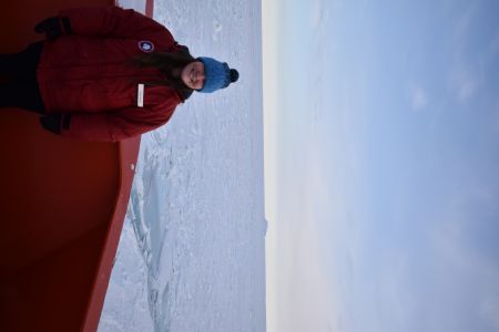 Lydia Hayes-Gaustella stands on a research vessel in winter wear with the solid, flat, white landscape of Antarctica in the background